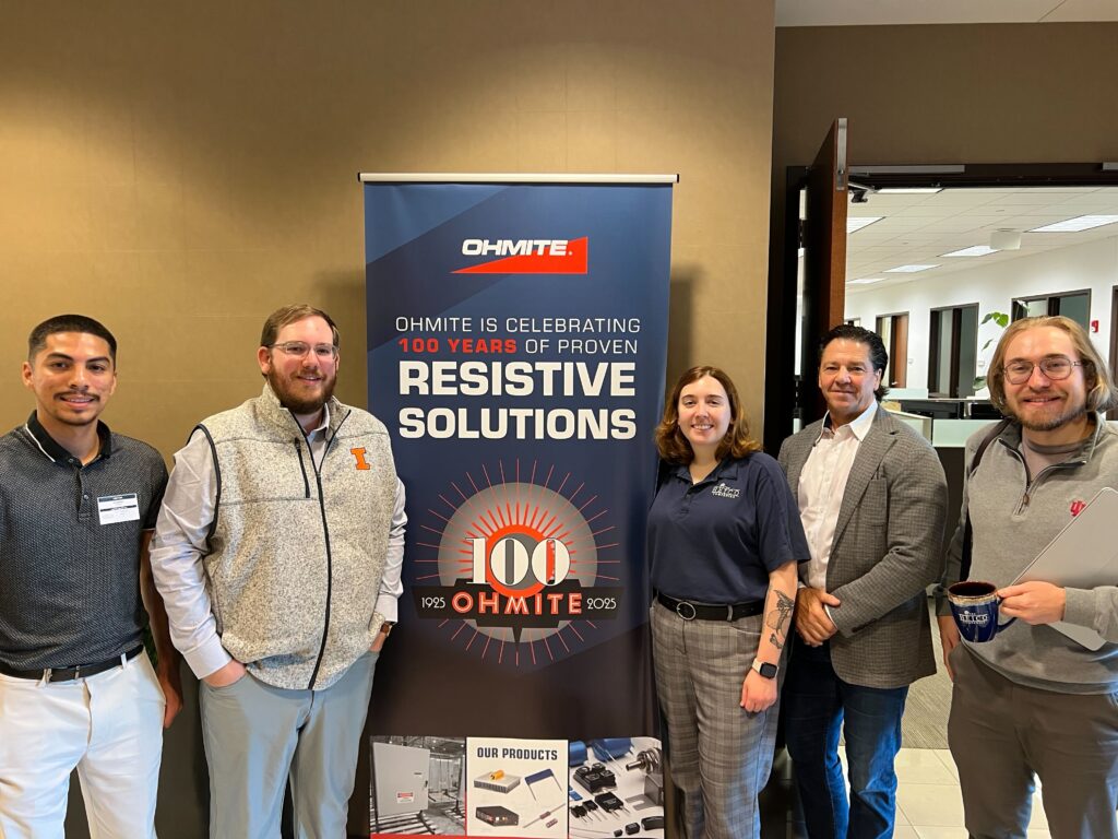 Five people stand beside an Ohmite banner for chassis mounted resistive solutions during an OuterBox client visit.