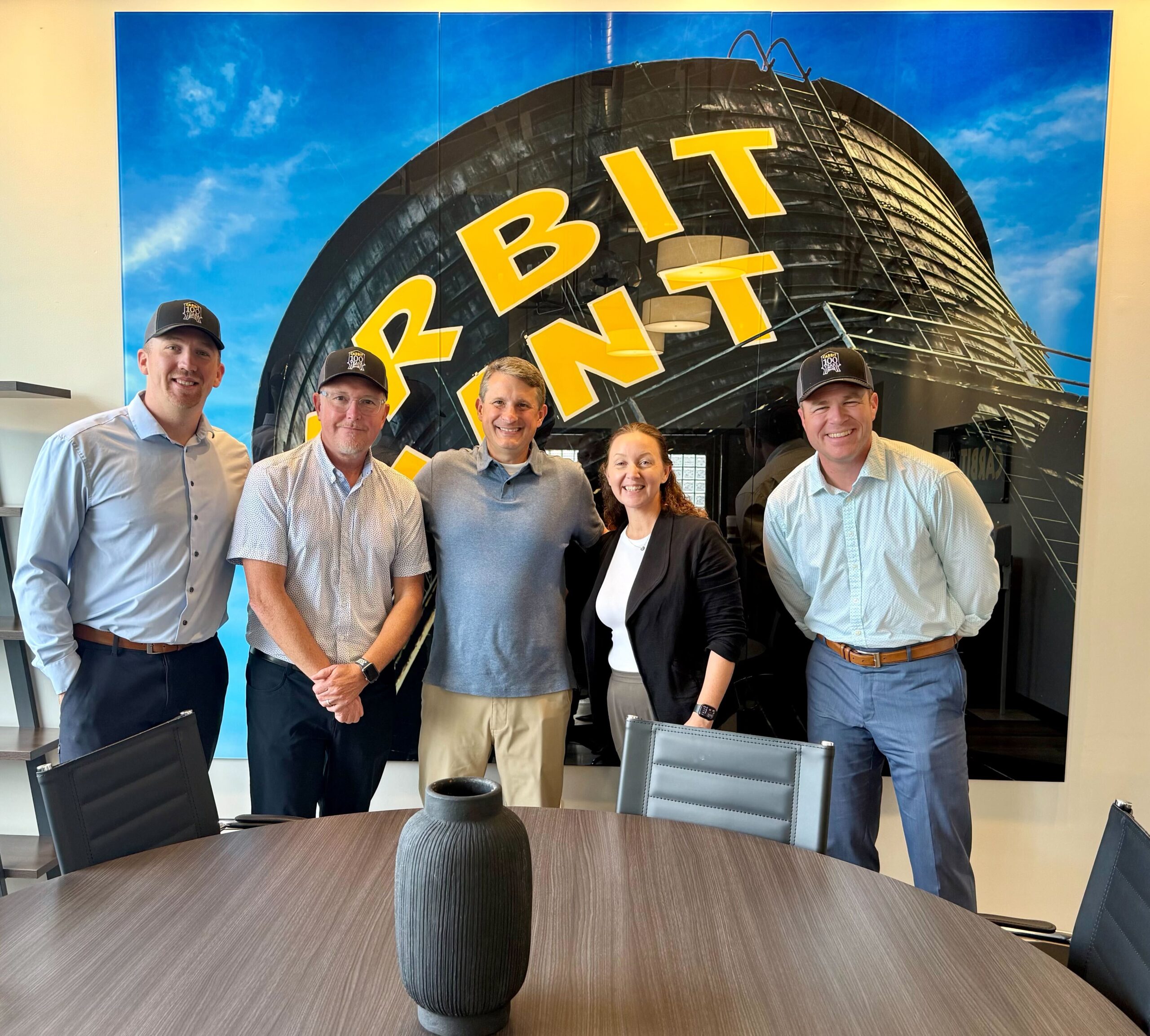 Six people stand in a conference room with a Carbit Paint wall graphic during an OuterBox client visit.