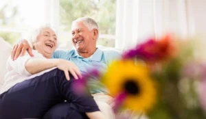 Smiling older couple relaxing together on a sofa in a bright, sunlit senior-living residence with fresh flowers in the foreground
